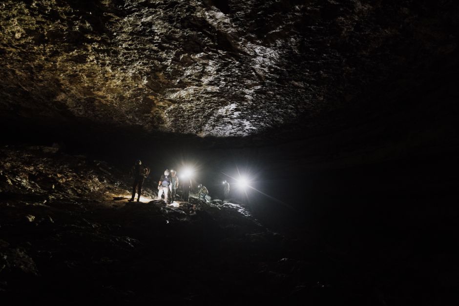 A team of spelunkers explore a dark cave with headlamps illuminating rocky cave formations.